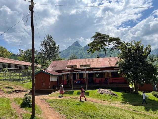 School building in a green field, with mountains in the background. Children walk around outside.