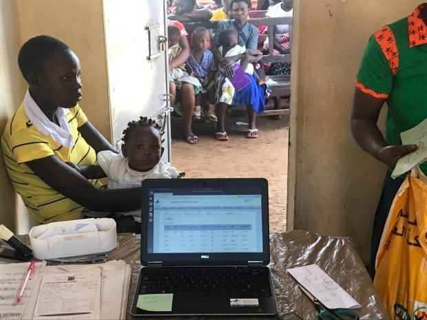 Woman with child at a laptop, with other children in the background. Indoors, possibly a clinic setting.