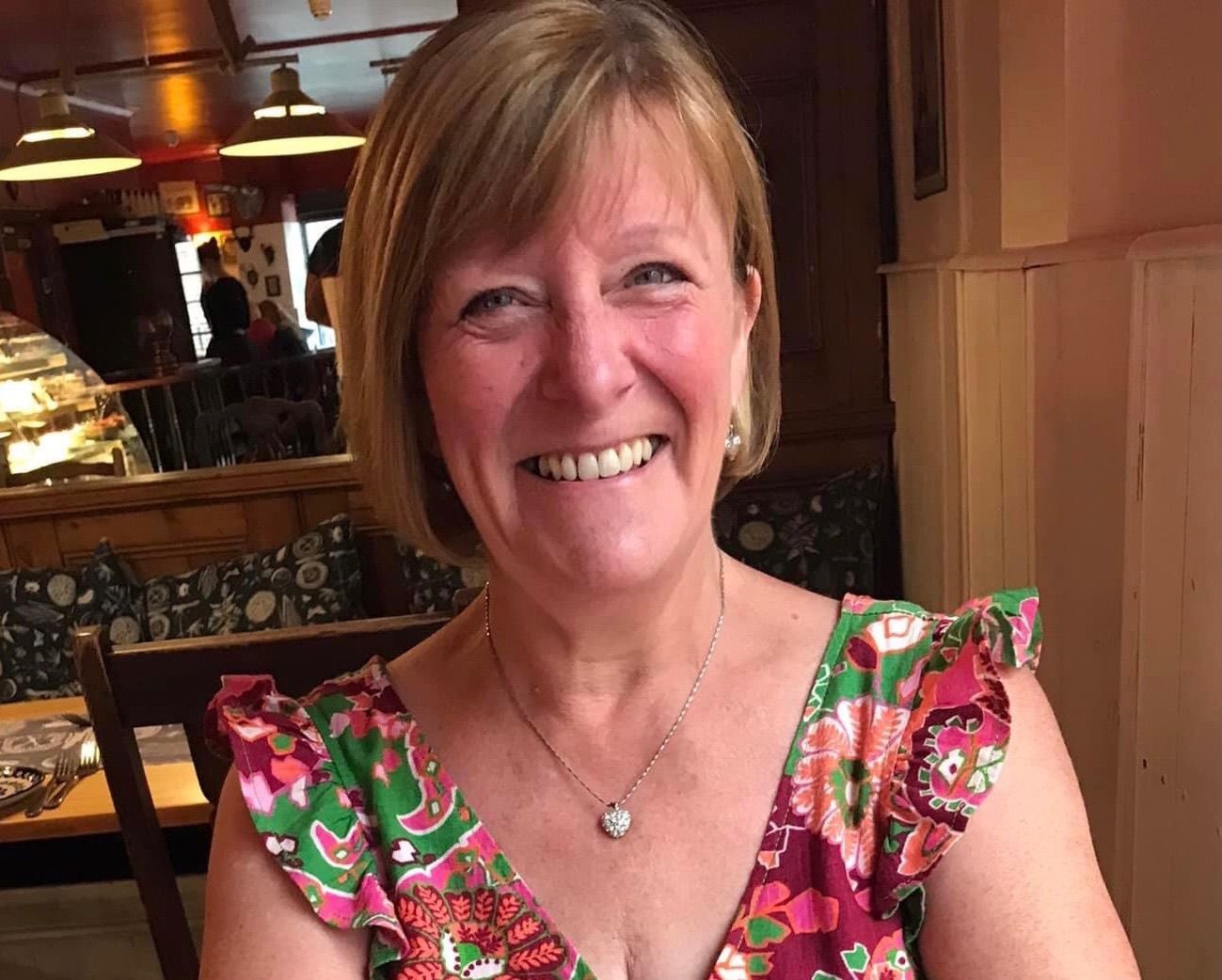 Woman with blonde hair smiles at the camera, wearing a floral top, seated in a restaurant.