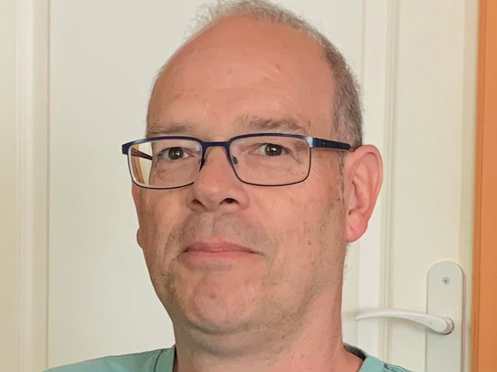 Man with glasses, looking at the camera, against a background of a white door and orange wall.