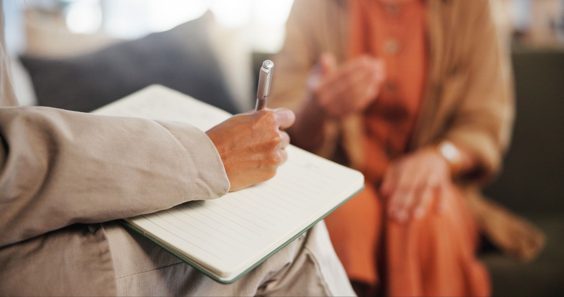 A person in a light suit writes in a notepad while another person gestures, blurred in the background.