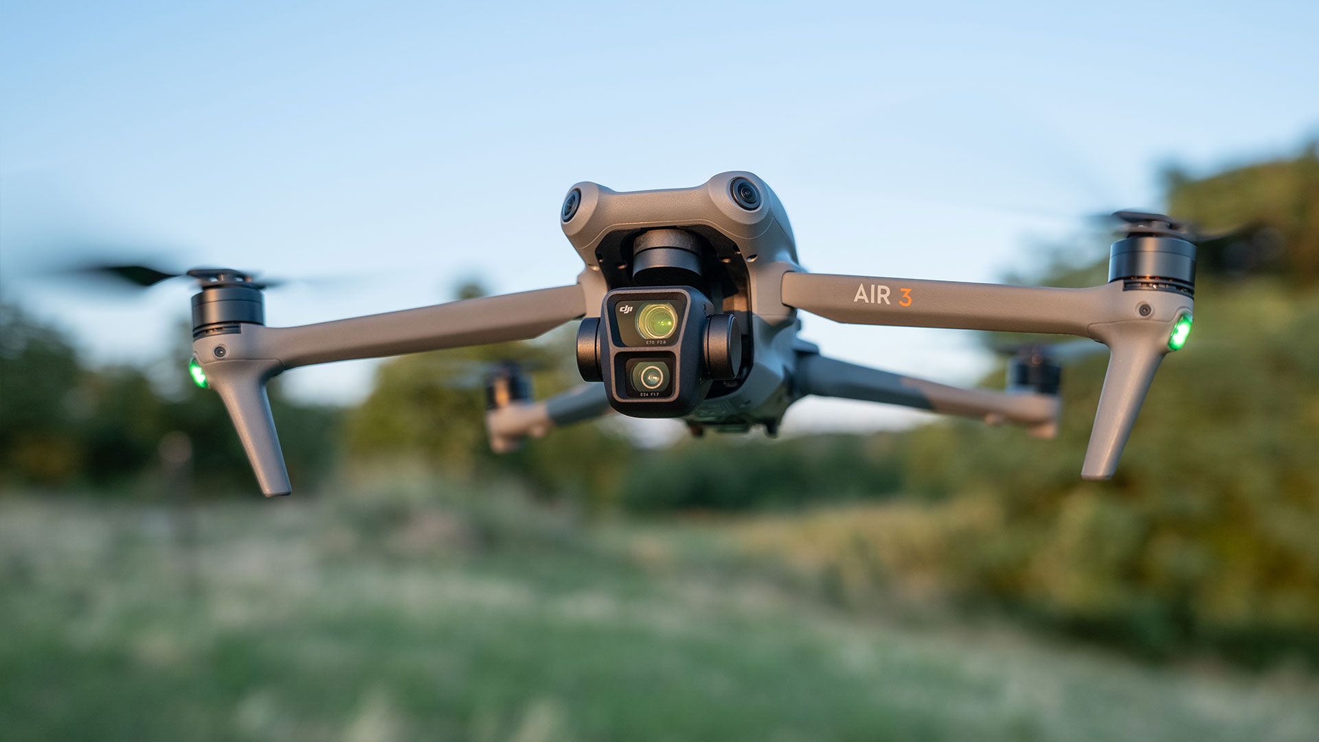 Gray drone in flight, facing camera, with two visible rotors turning against a blurred outdoor background.