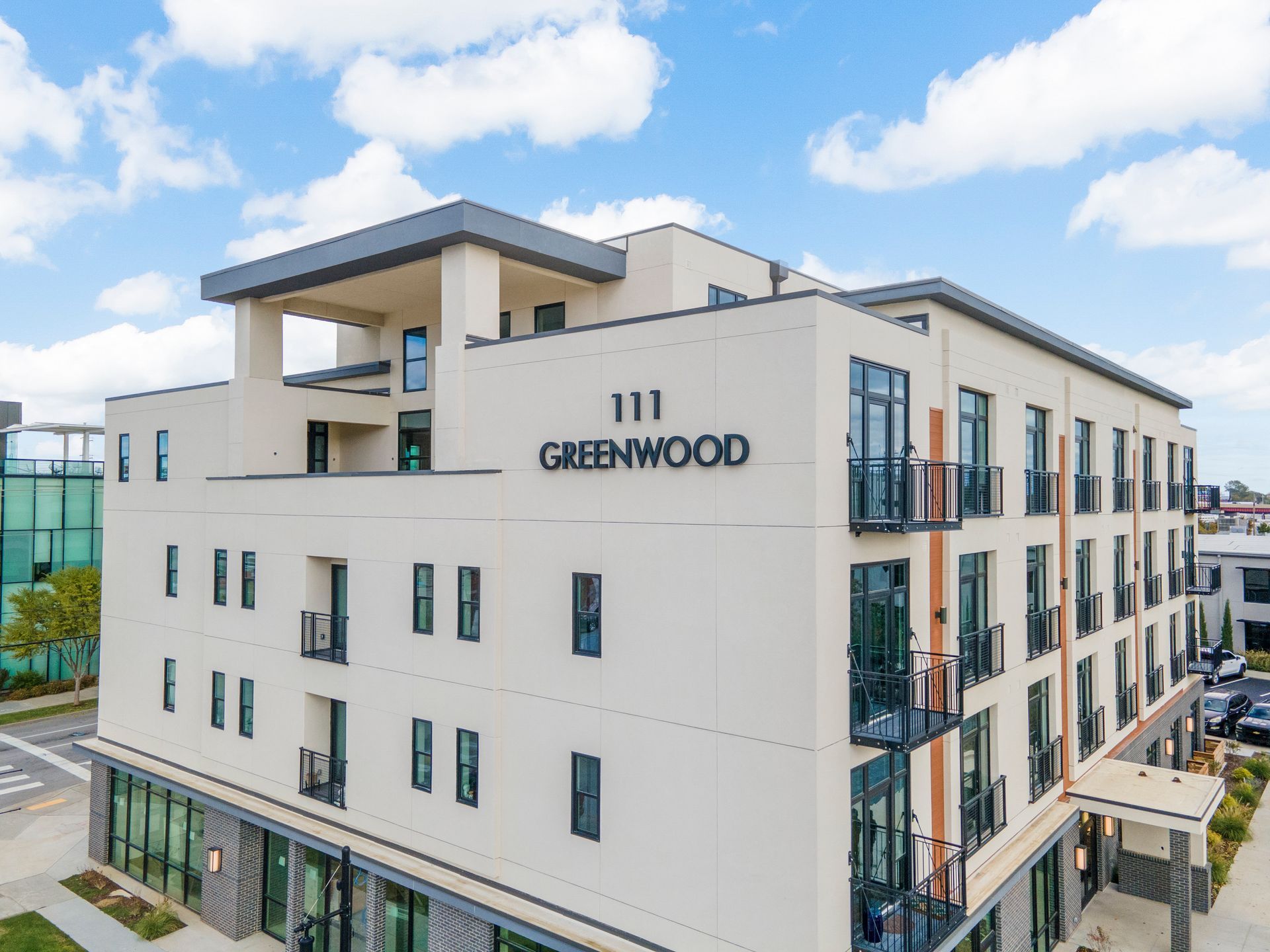 Exterior of a modern apartment building, 111 Greenwood, with beige facade, balconies, and retail space on the ground floor.