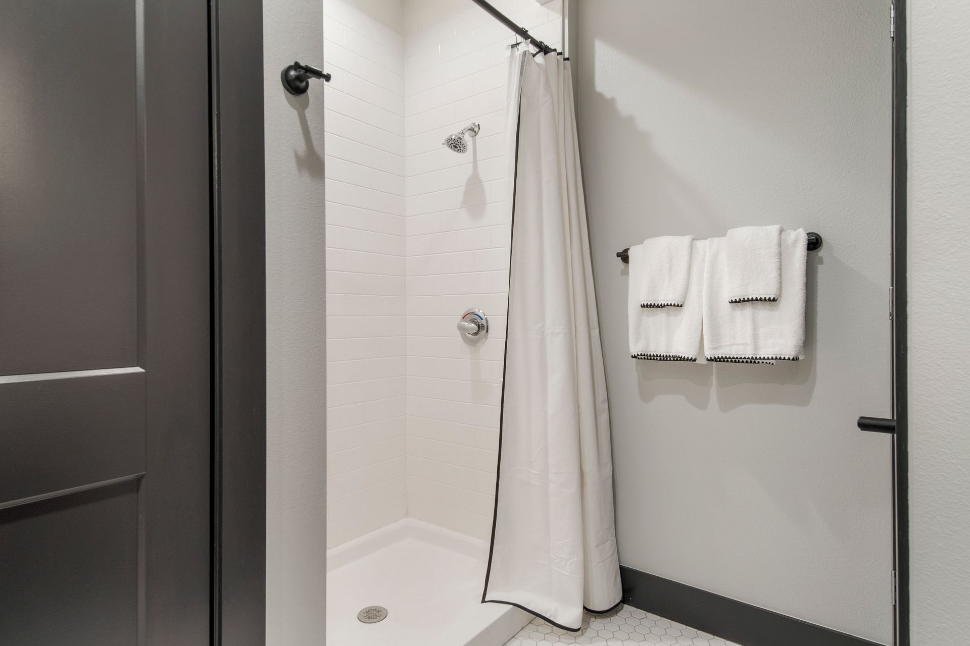 Shower with white tile walls and a white shower curtain with black trim. Towel bar with towels.