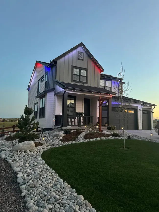 Two-story house with white siding and gray roof, lit with red and blue lights; landscaped yard.