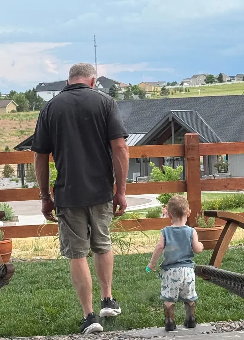 Man and small child walk on grass toward a wooden fence. Houses and hills in the background under a cloudy sky.