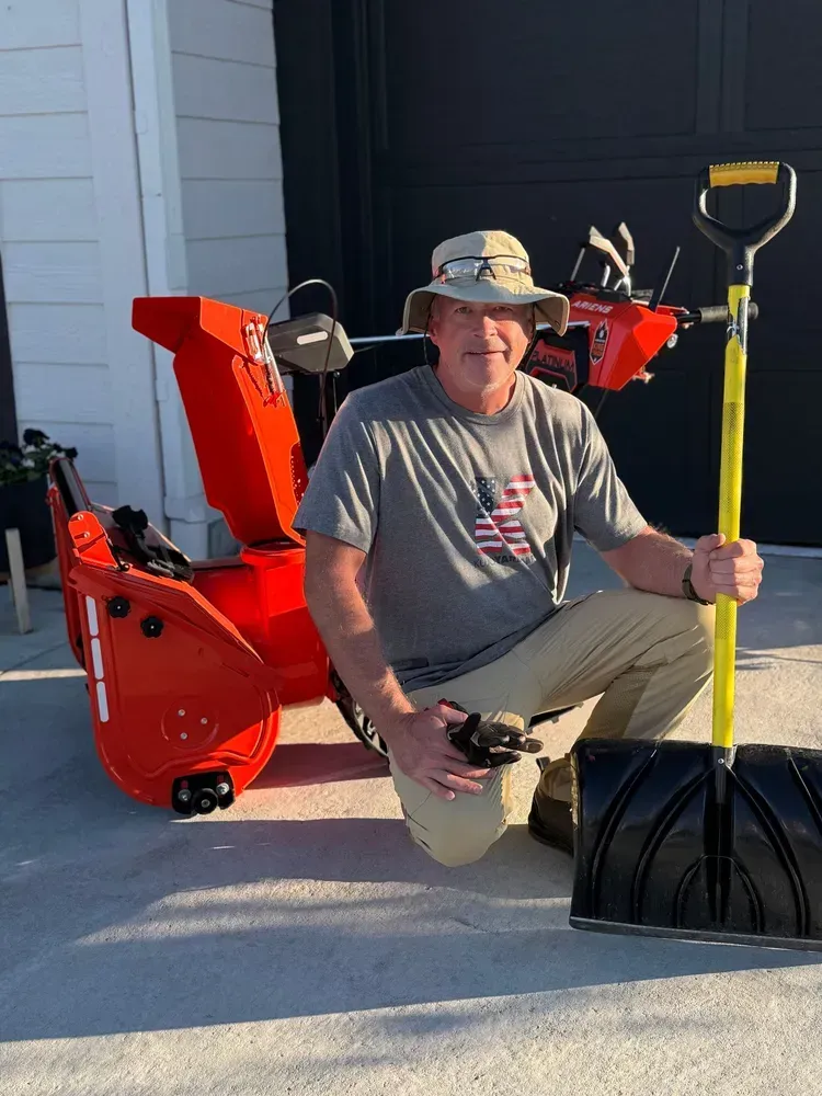 Man kneels next to a snowblower and shovel. He wears a hat and holds the shovel. Orange snowblower in the background.