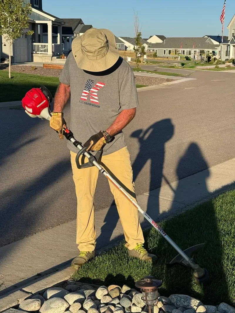 Person wearing a hat and gloves uses a string trimmer on grass near a sidewalk.