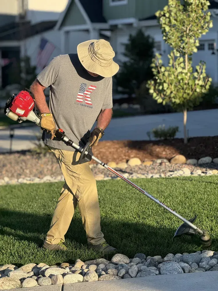 Man in a hat using a string trimmer on a lawn, trimming near rocks, outside a house.