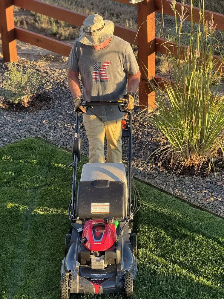 Person mowing a green lawn with a push mower; wearing a hat, gloves, and khaki pants.