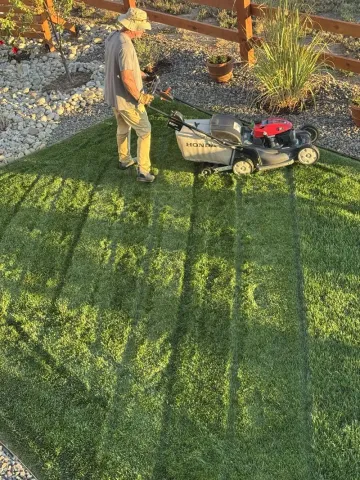 Man mowing a lawn with a red and black lawnmower in a yard with a wooden fence.