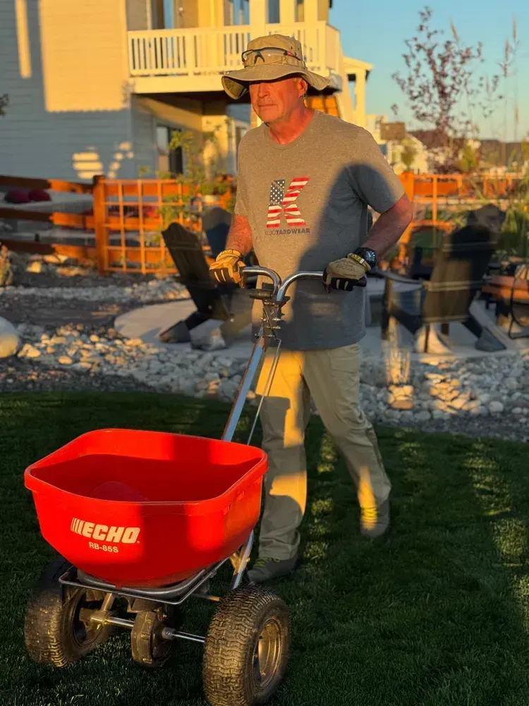 Man pushing a red fertilizer spreader on a lawn, wearing a hat, gloves, and a shirt with an American flag design.