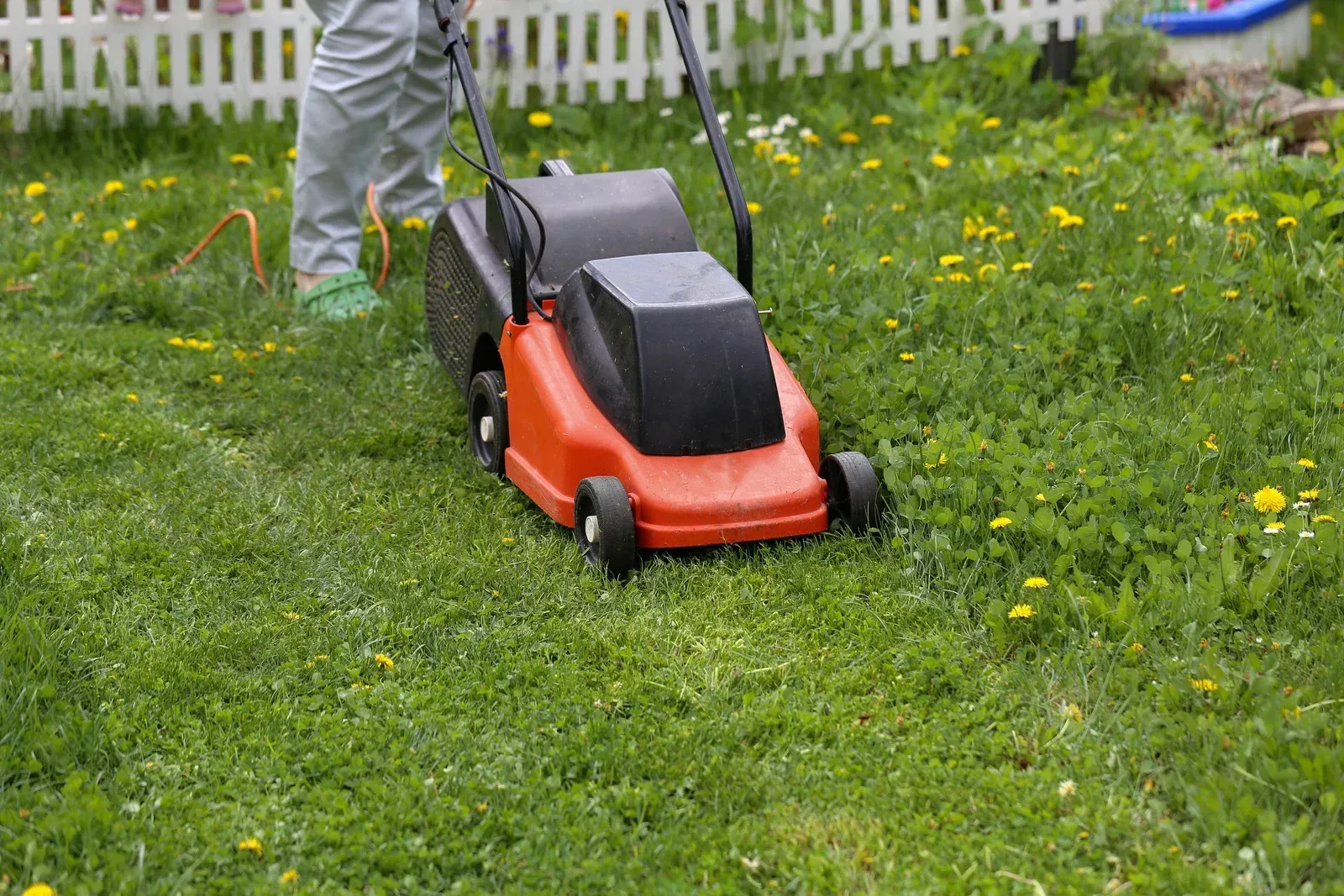 Person mowing a grassy lawn with a red and black lawnmower near a white fence.