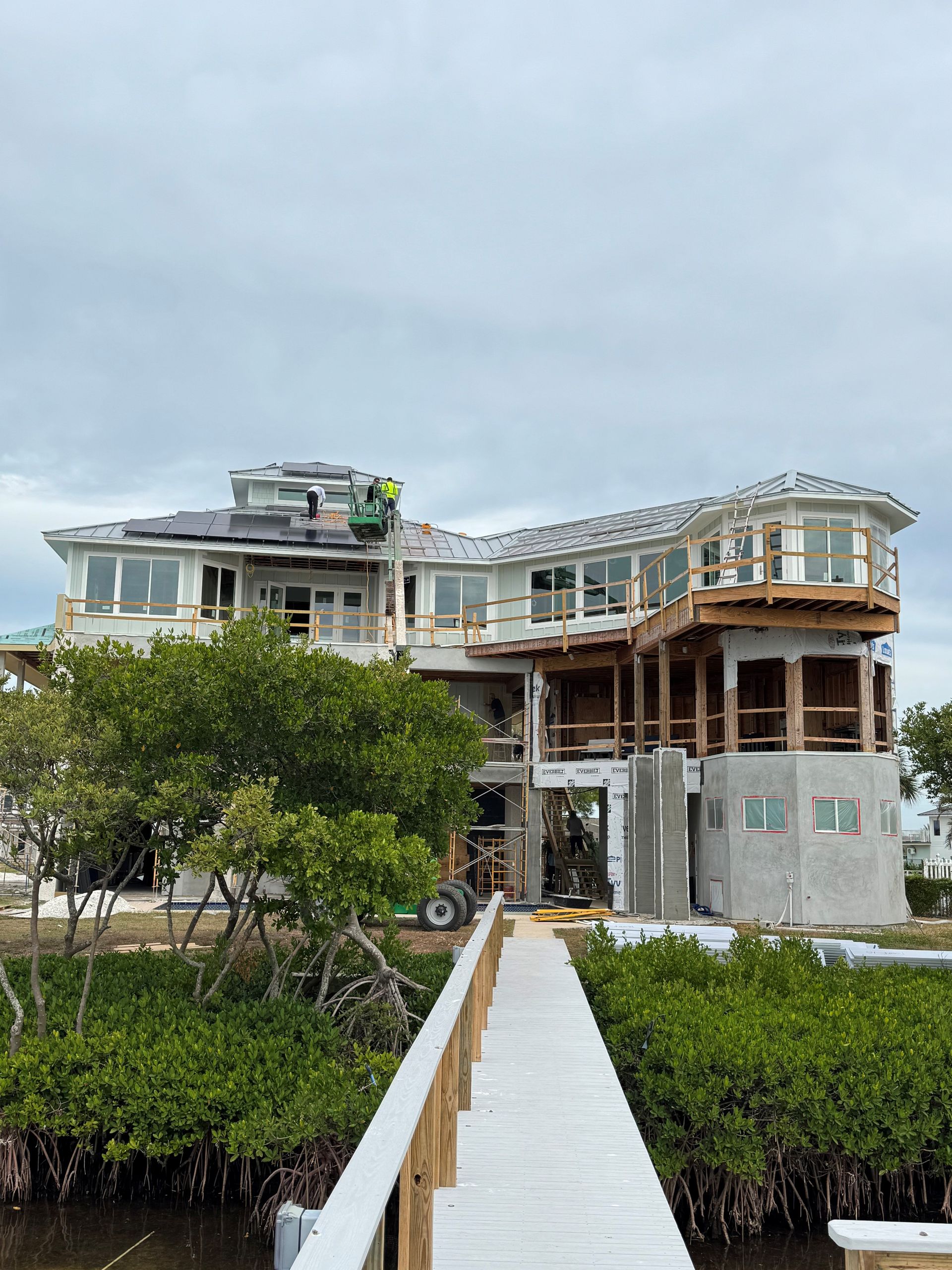 Partially built white house with scaffolding beside a narrow boardwalk over mangroves under cloudy sky