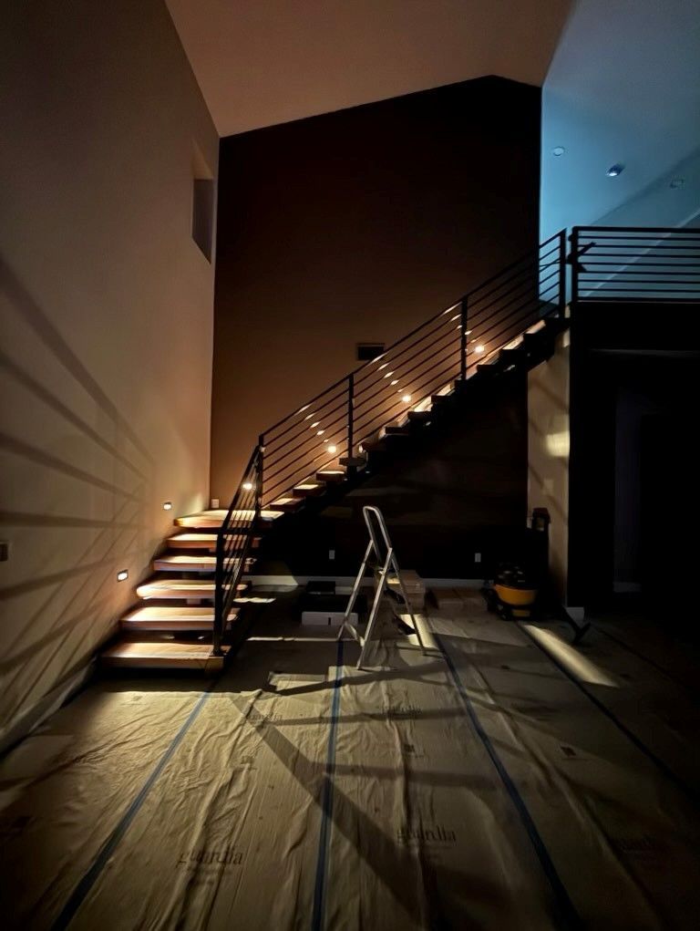Dim staircase with illuminated steps, metal railing, and a ladder in a dark interior under renovation