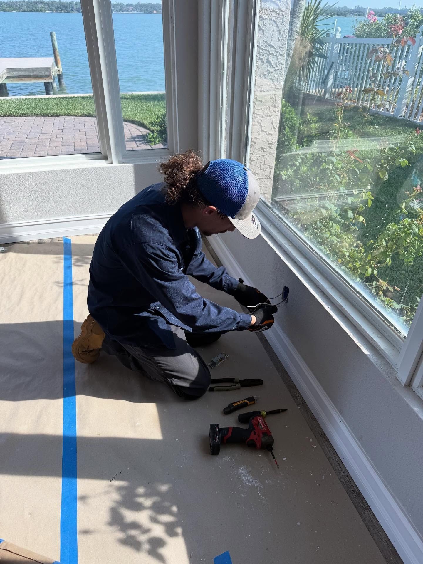 Electrician kneeling by a window, working with tools on the baseboard in a bright room by the water.