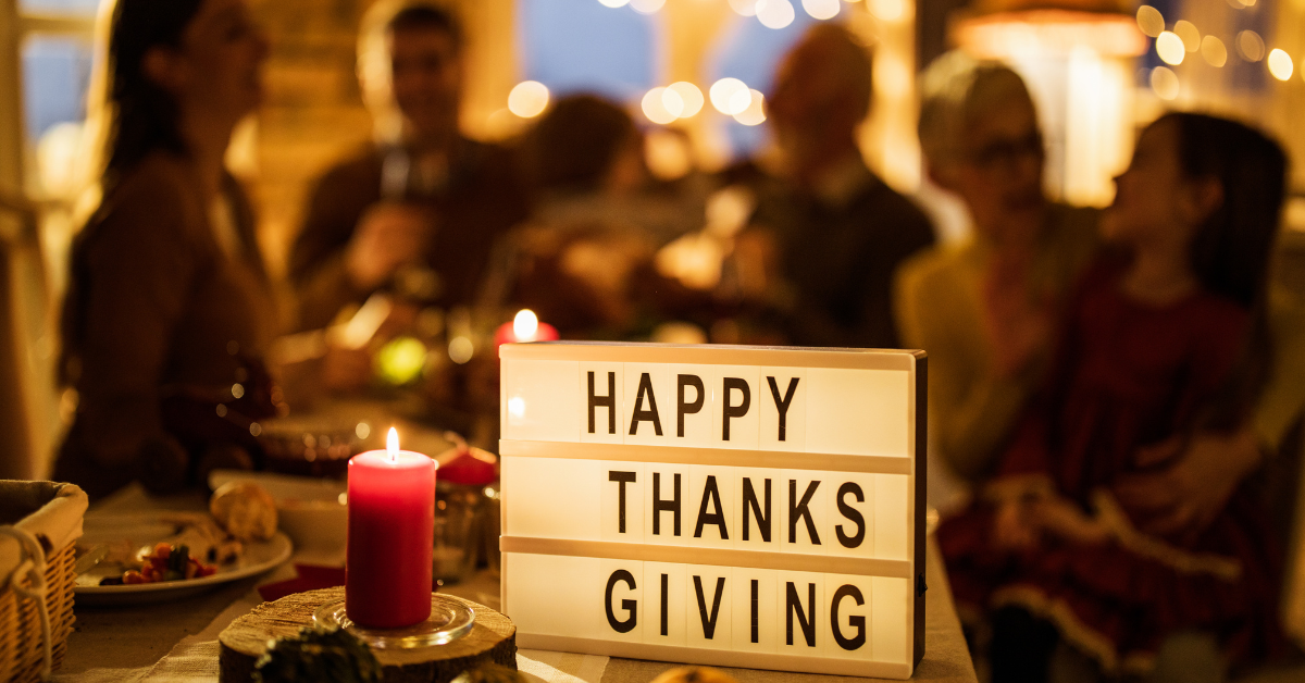 Thanksgiving table with a lit candle and sign reading “Happy Thanksgiving,” with people blurred in the background