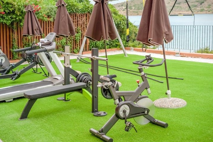 Outdoor gym area with fitness equipment including a treadmill and stationary bike on green turf near a waterfront.