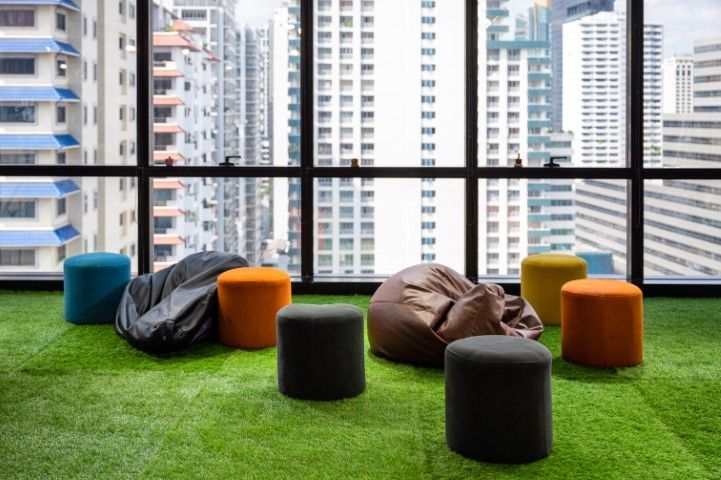 Colorful stools and beanbag chairs sit on a green turf floor in front of a window overlooking a city skyline.