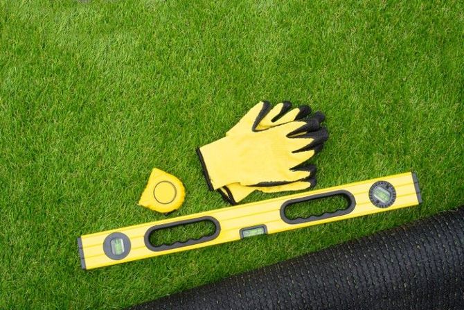Yellow work gloves, a tape measure, and a spirit level placed on a patch of artificial grass next to a roll of turf.