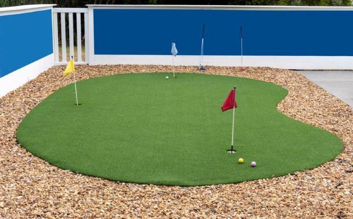 A backyard putting green with three flags and two golf balls on artificial grass surrounded by gravel.