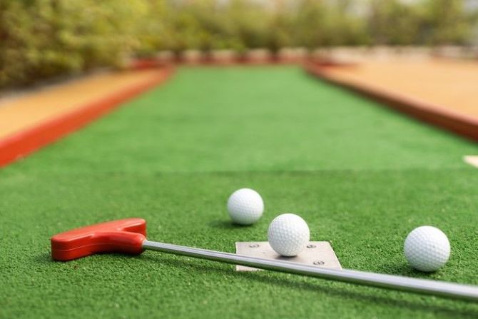 A red mini-golf putter rests on a green artificial turf course with three white golf balls nearby.