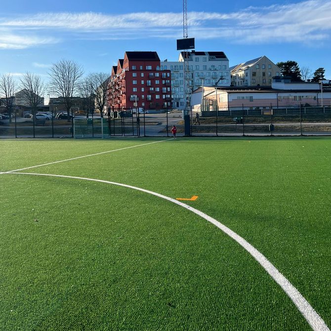 A green artificial turf field with a white curved line, set against a background of urban buildings under a blue sky.