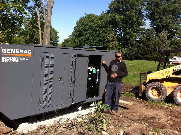 Man standing beside a gray Generac industrial generator outdoors near a yellow skid-steer loader.