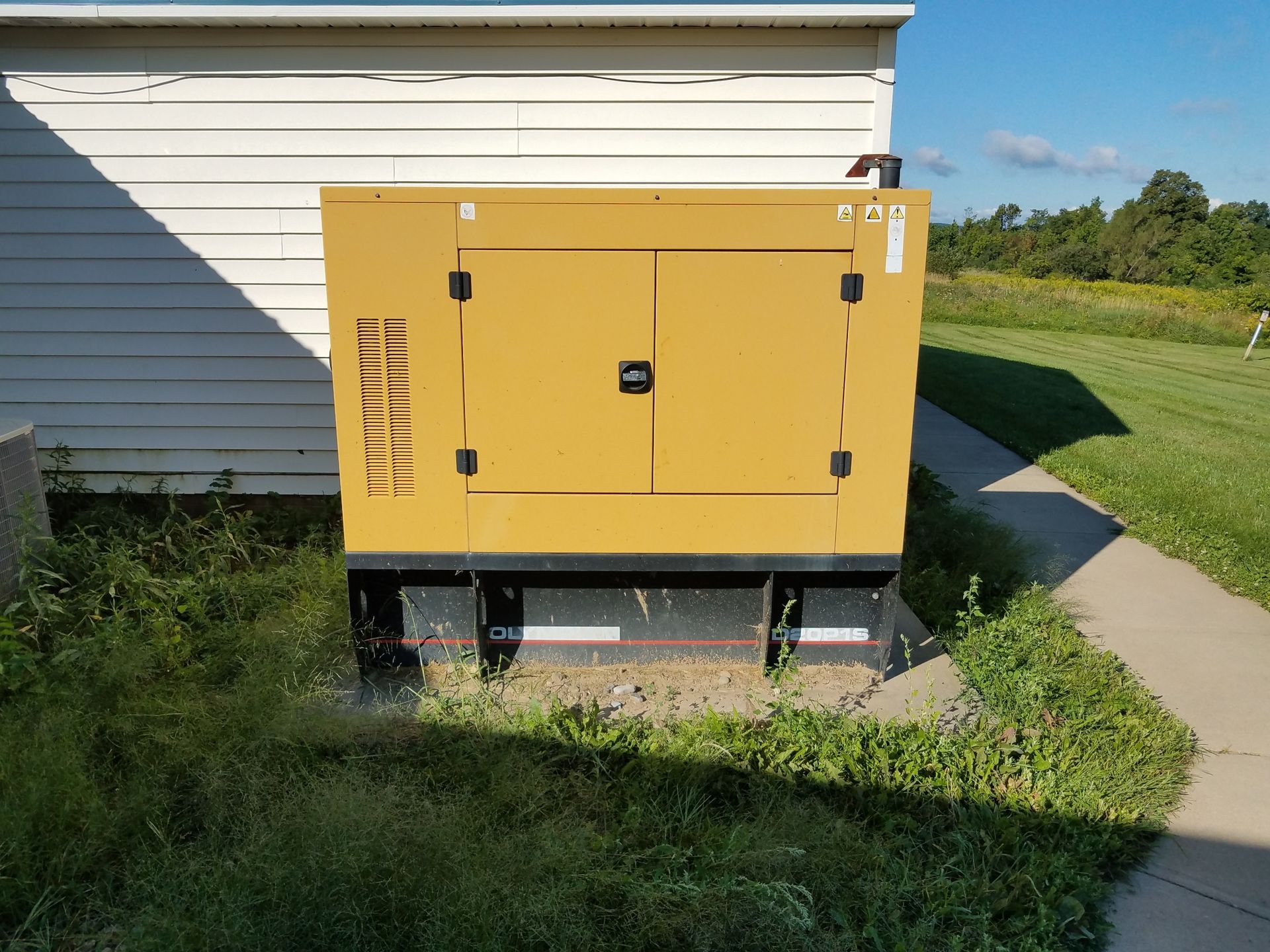 Yellow generator beside a white house, standing on a concrete pad in a grassy yard.
