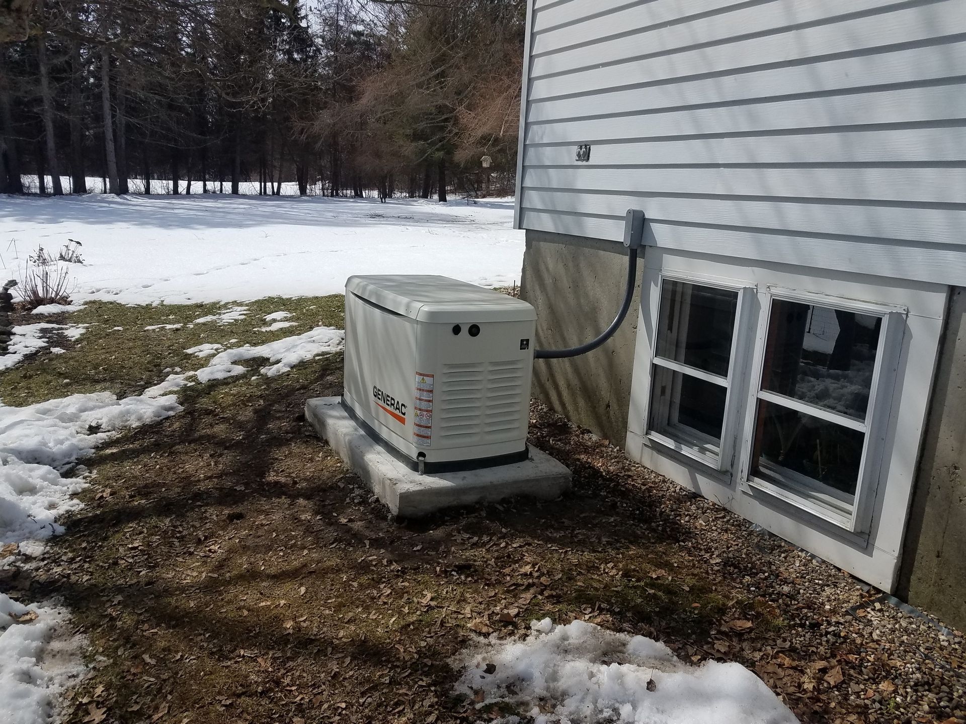 Generator beside a house on a snowy lawn, with a basement window and siding visible
