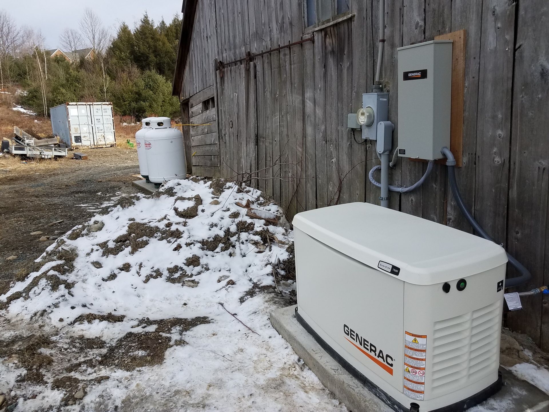Outdoor generator and electrical equipment beside a weathered wooden building with patchy snow on the ground