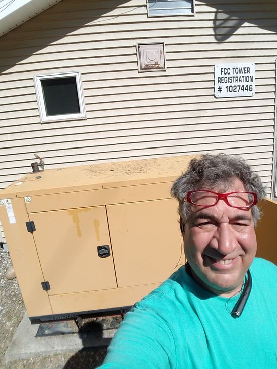 Man smiling outdoors beside a beige generator against a siding wall, wearing a teal shirt and red sunglasses on his head