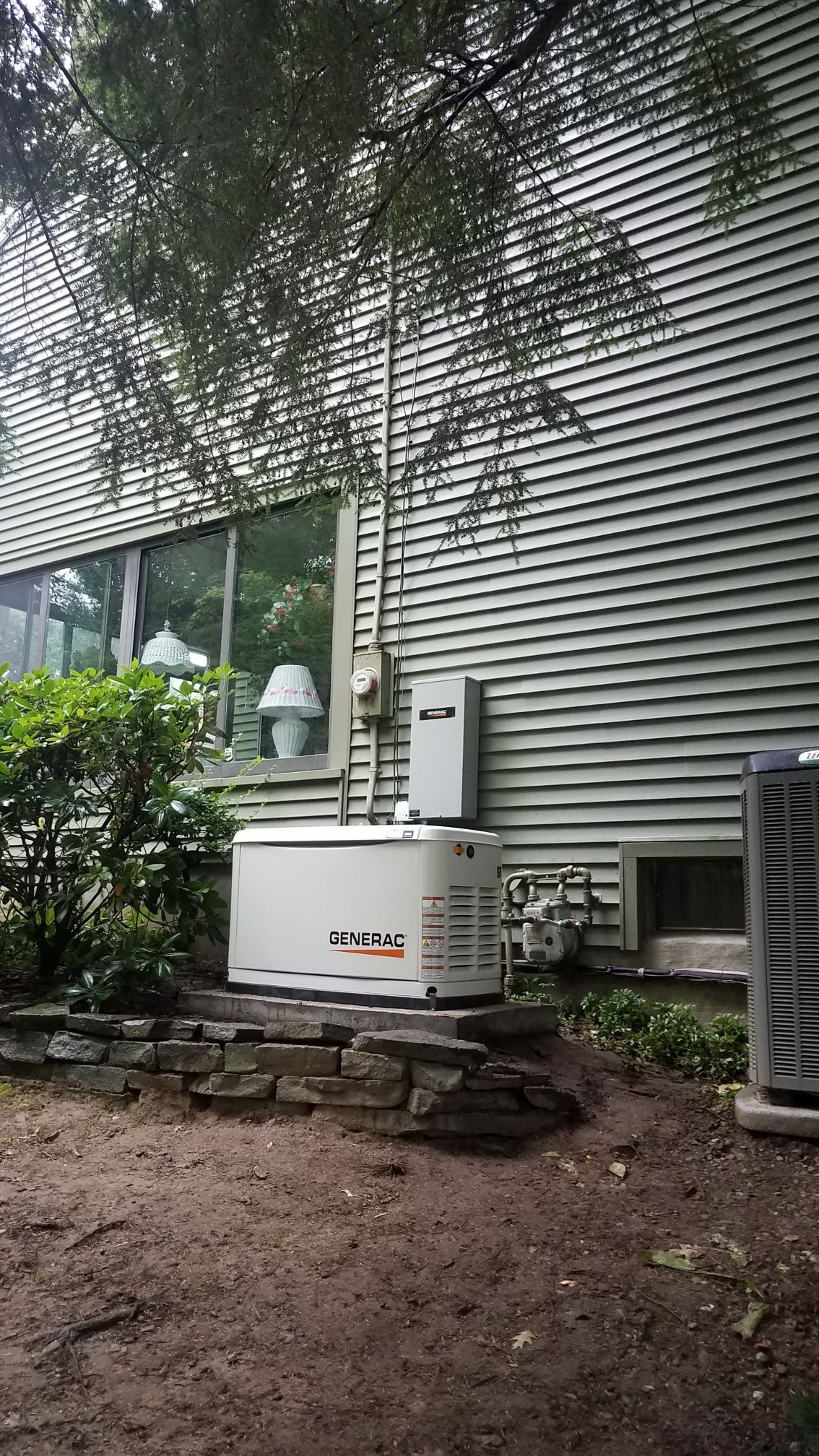 Outdoor HVAC unit beside a gray building with a window, stone edging, and muddy ground below.