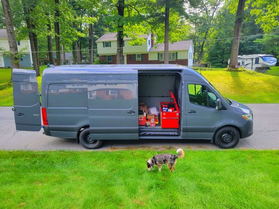 Gray van with side door open showing supplies, parked by a grassy yard with a small dog in front.