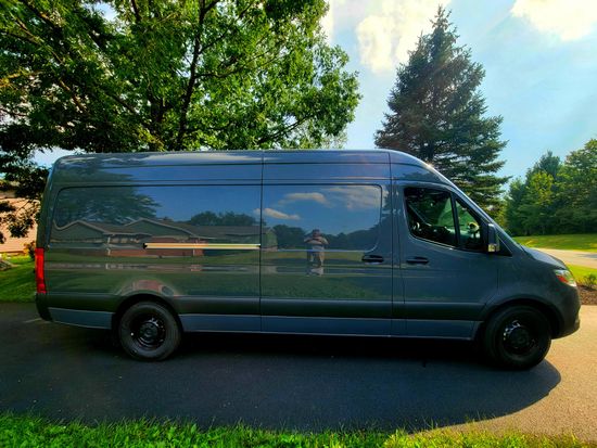 Dark gray cargo van parked on a road beside trees in a sunny park-like setting