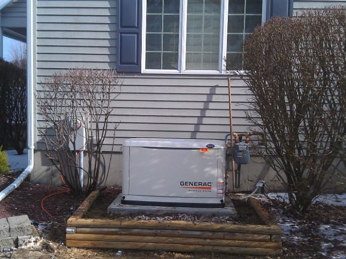 Outdoor generator beside a house, surrounded by shrubs and wooden planks, with light snow on the ground.