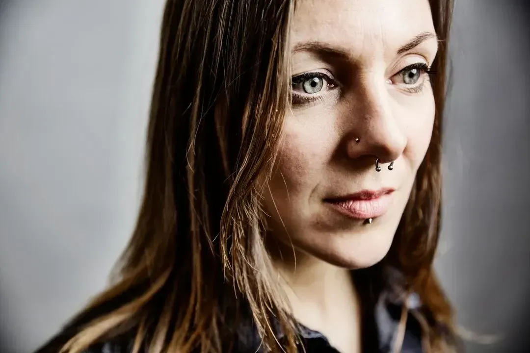 Close-up portrait of a woman with brown hair and a nose ring, looking off to the side against a blurred gray background
