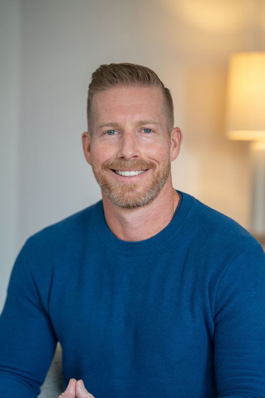 Smiling man in a blue shirt standing indoors beside a lit lamp