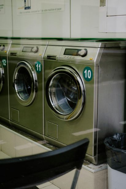 A row of stainless steel washing machines in a laundromat, numbered 10, 11, and 12, next to a black chair.