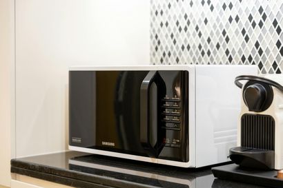 A white and black microwave sits on a black counter next to a coffee machine, against a patterned diamond tile backsplash.