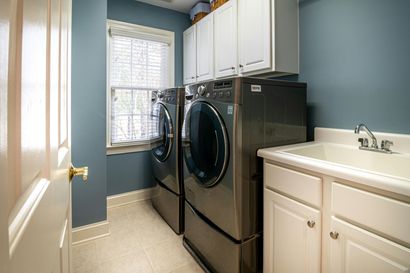 A laundry room with blue walls, white cabinets, a utility sink, and two dark gray front-loading machines by a window.