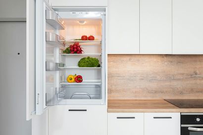 A modern kitchen with a built-in refrigerator open, stocked with fresh vegetables on white shelves.
