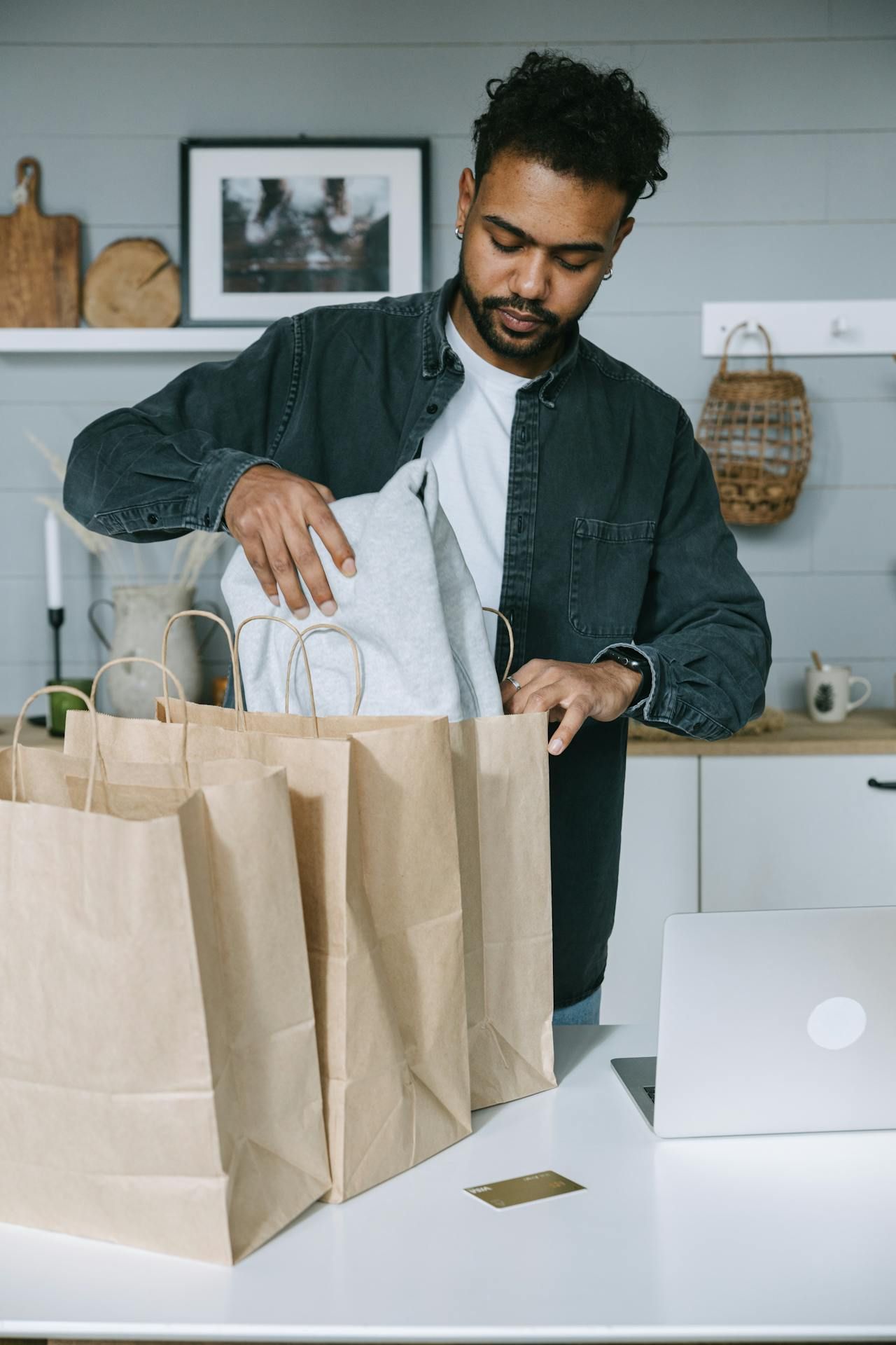 A person in a dark green shirt unpacking white items from brown paper bags on a table next to a laptop and credit card.
