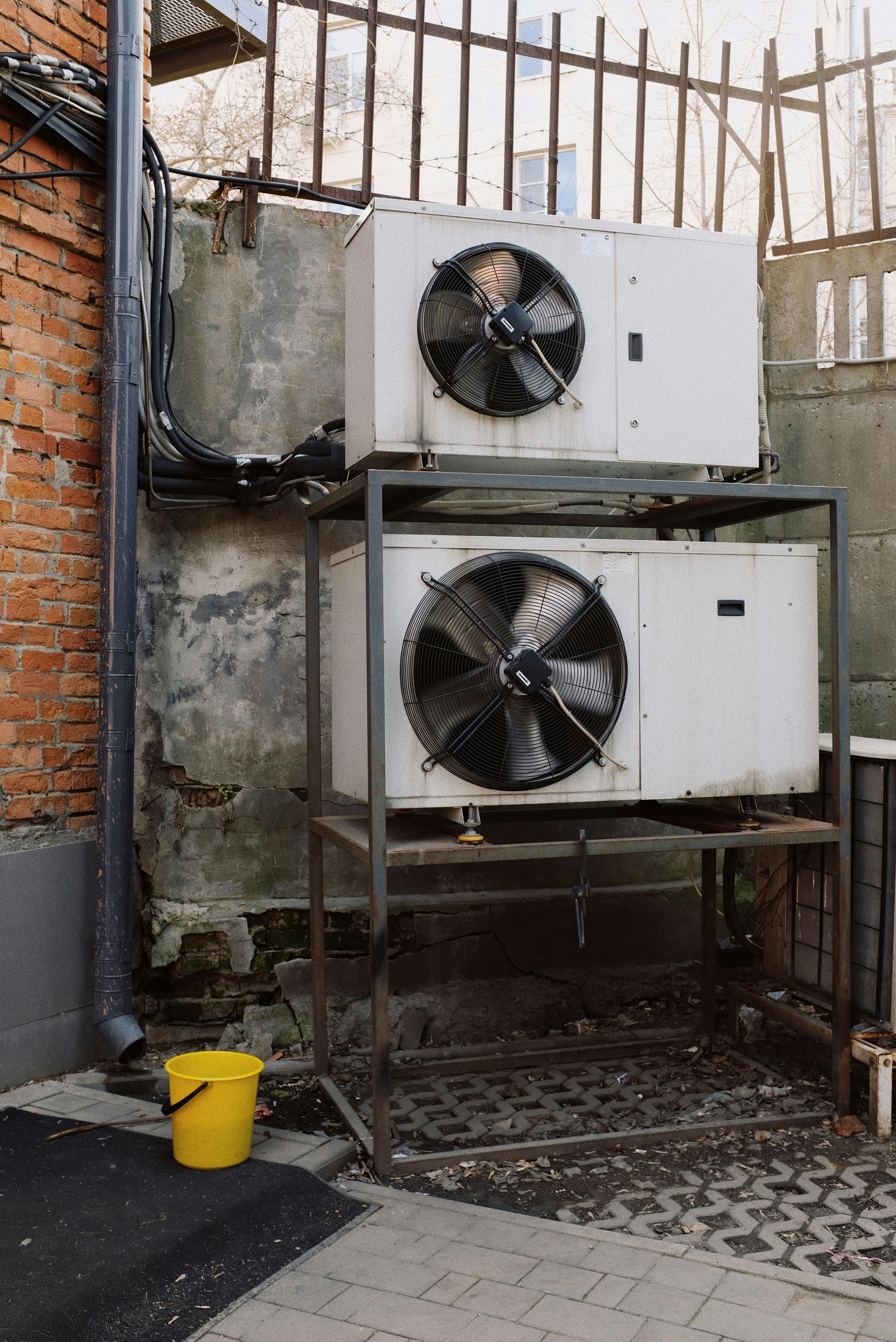 Two white air conditioning units stacked, with fans, on a metal frame, against a concrete wall.