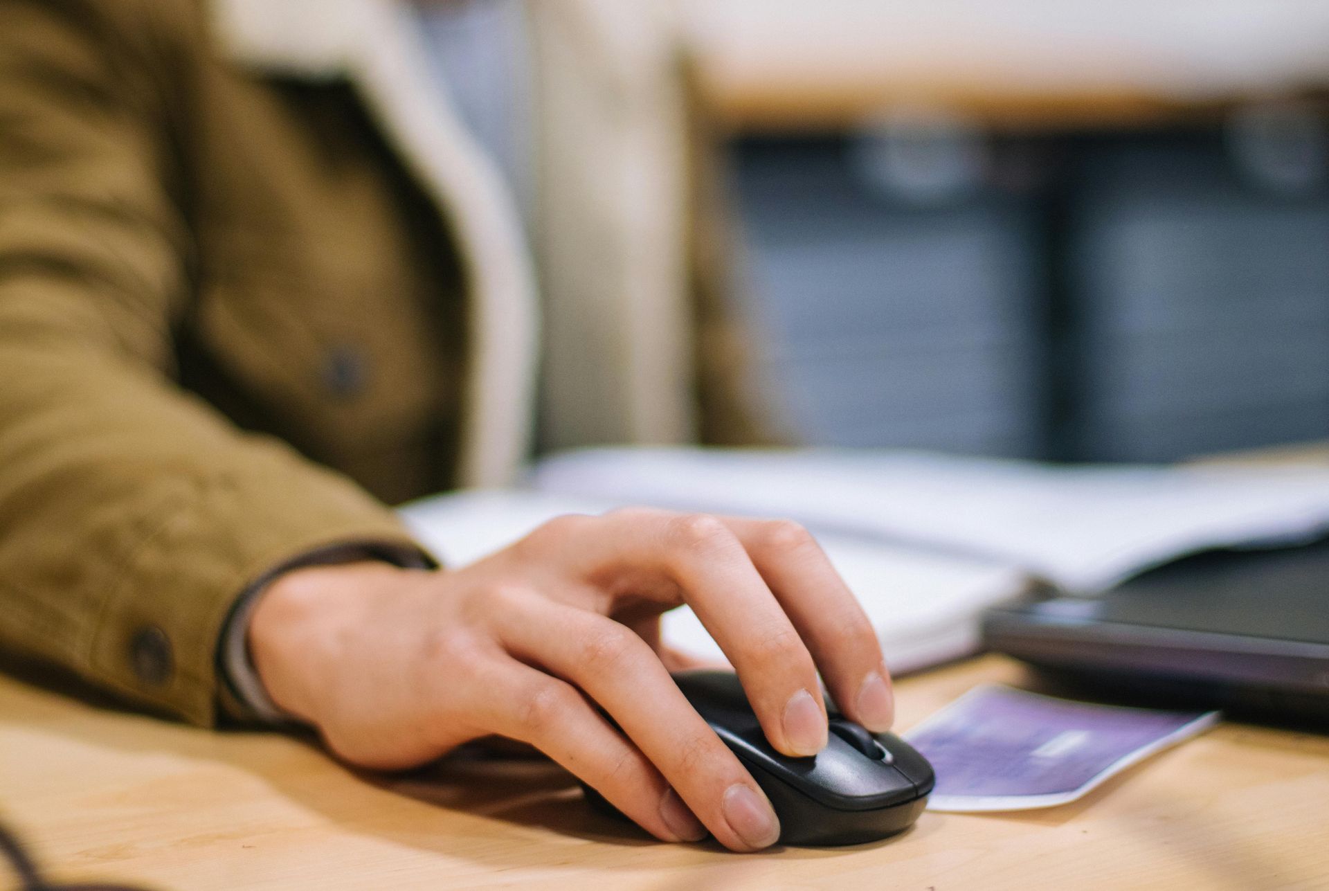 Hand using a black wireless mouse at a desk beside a laptop and notebook