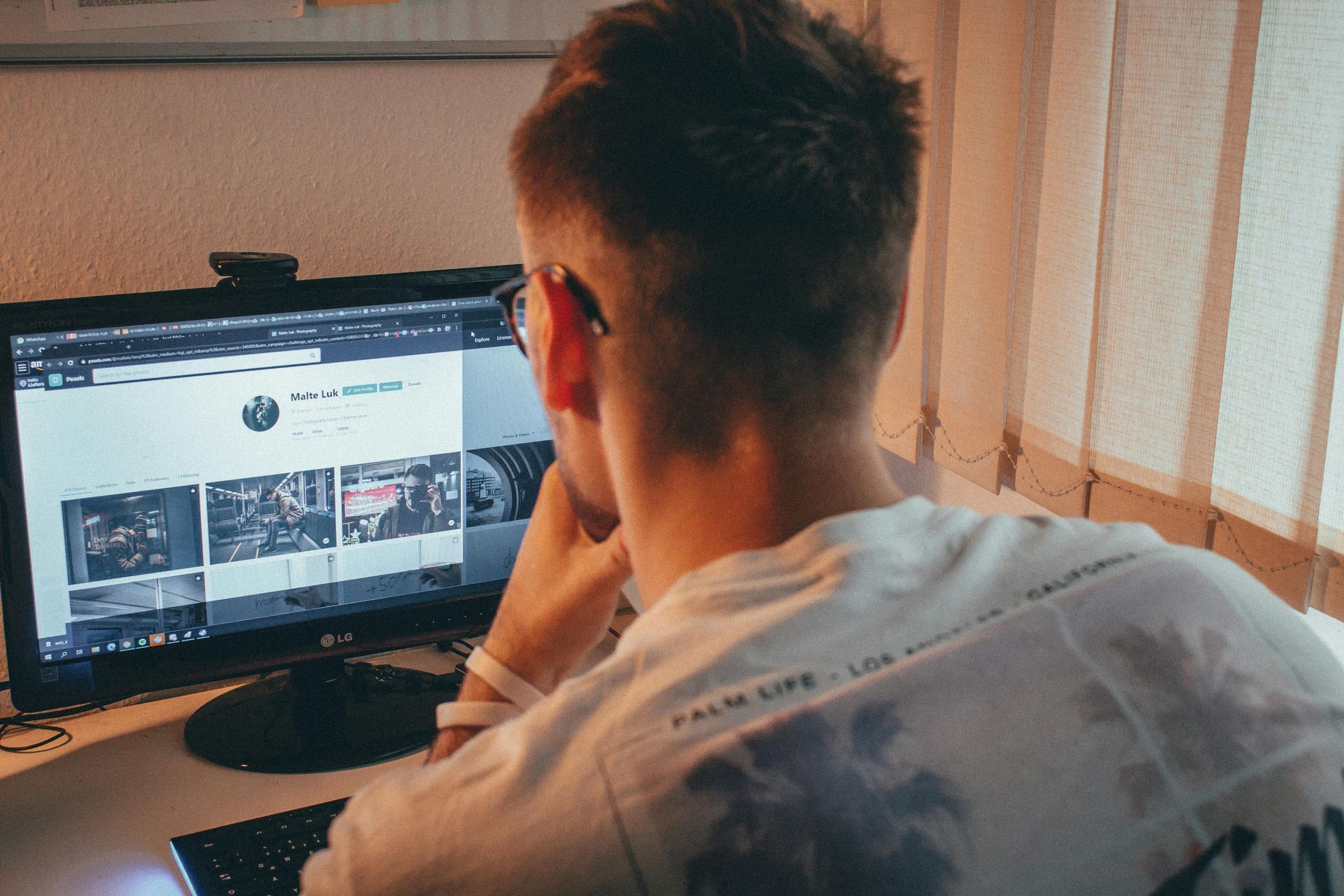 Person viewed from behind at a desk, looking at a computer monitor in a warm-lit room