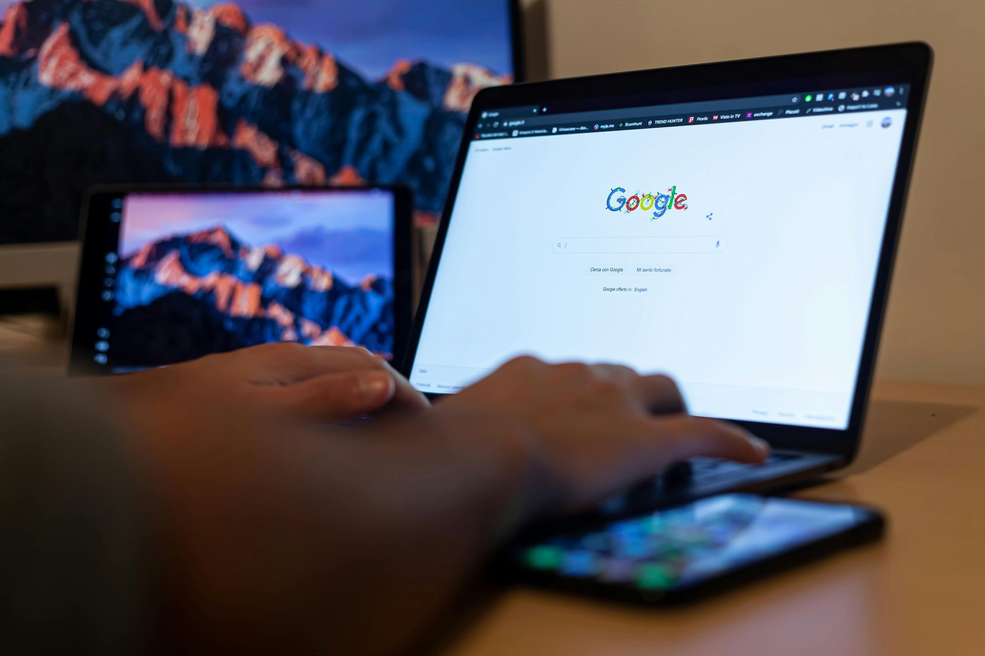 Person typing on a laptop with Google open, next to a smartphone on a desk.