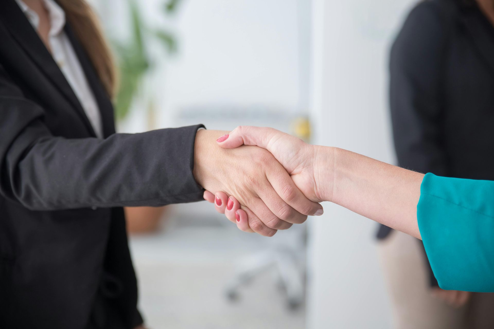 Two people shaking hands in a business setting, one in a black suit and one in a teal sleeve