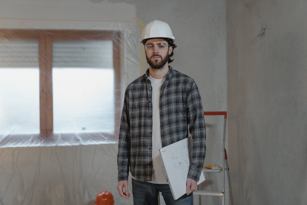 Contractor in a hard hat, plaid shirt, holding blueprints inside a room under renovation.