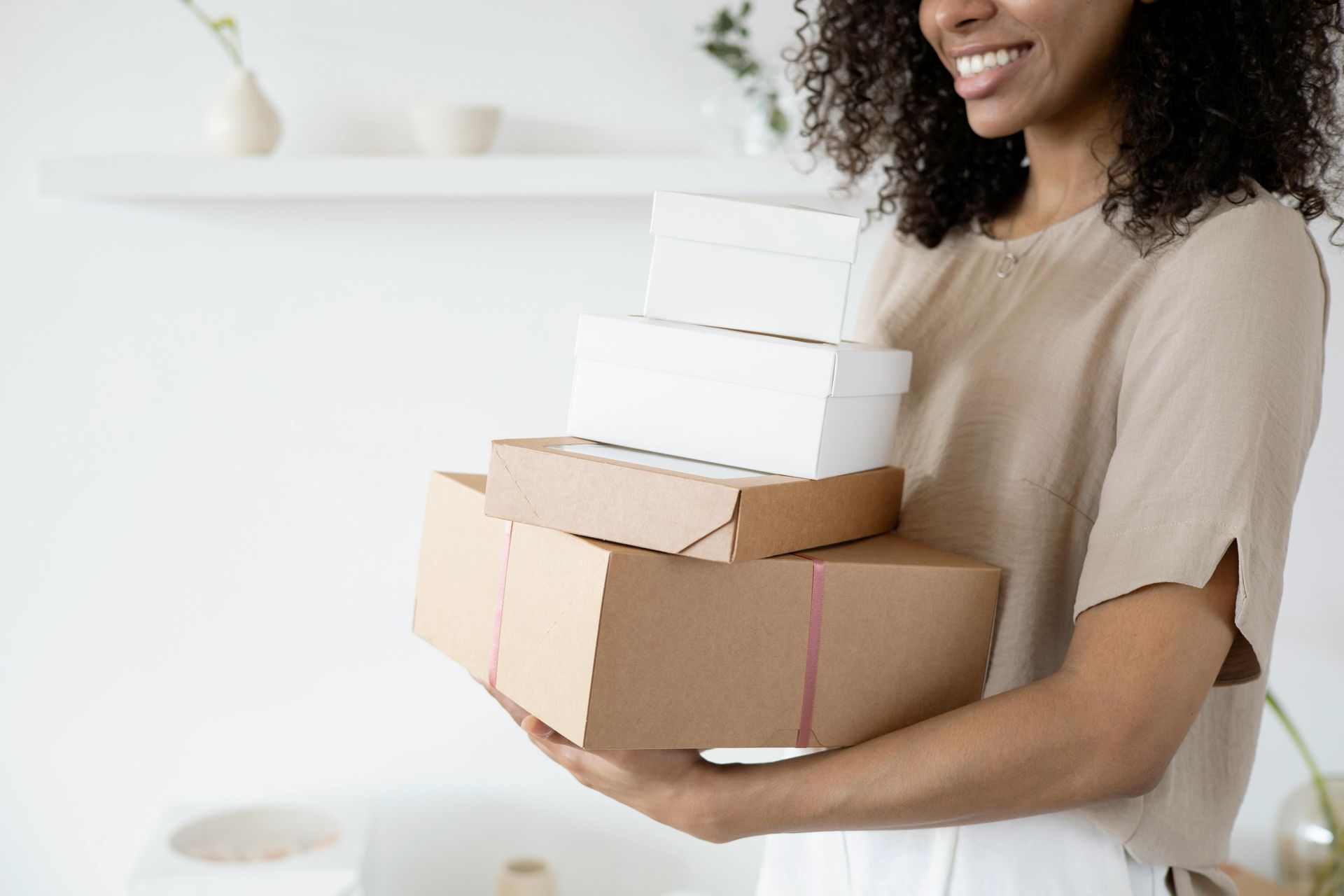 A smiling person holding a stack of four cardboard and white shipping boxes in a bright, modern room.